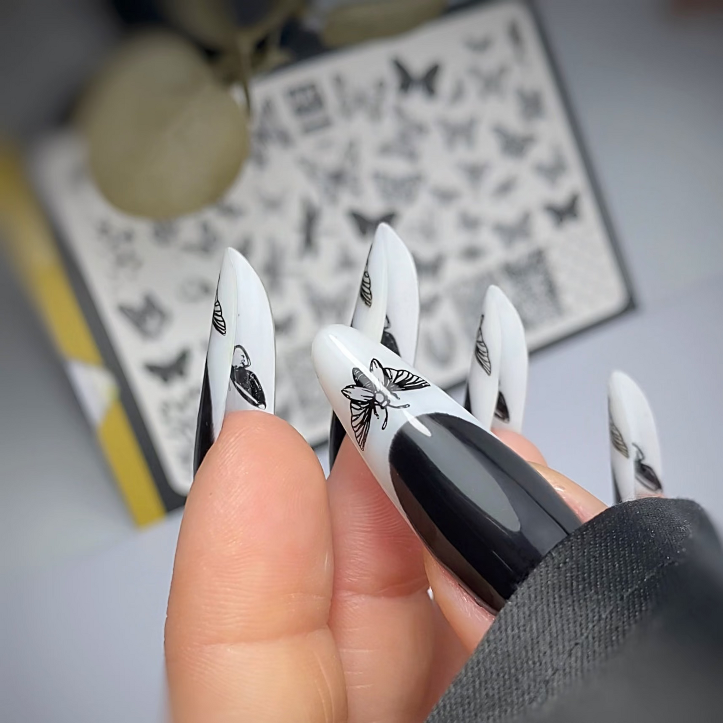 Black and white nail art designs held by a hand with a blurred background of butterfly stamping plates.