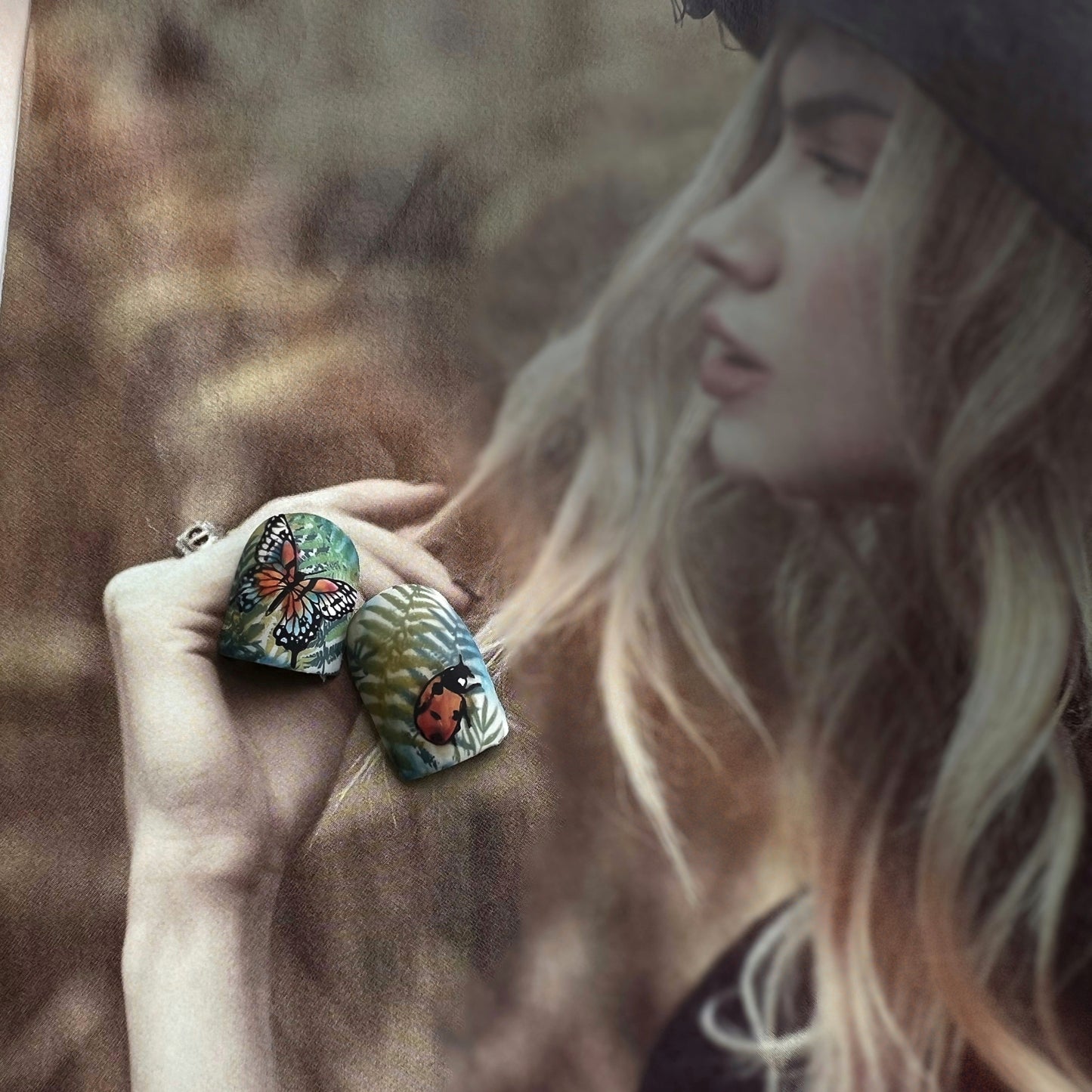 Two painted nail tips with butterfly and ladybug designs against a blurred magazine picture of a beautiful woman on background