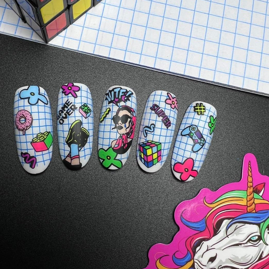 Decorative nail art with cartoon characters and symbols on a black surface with a Rubik's cube and grid paper in the background.