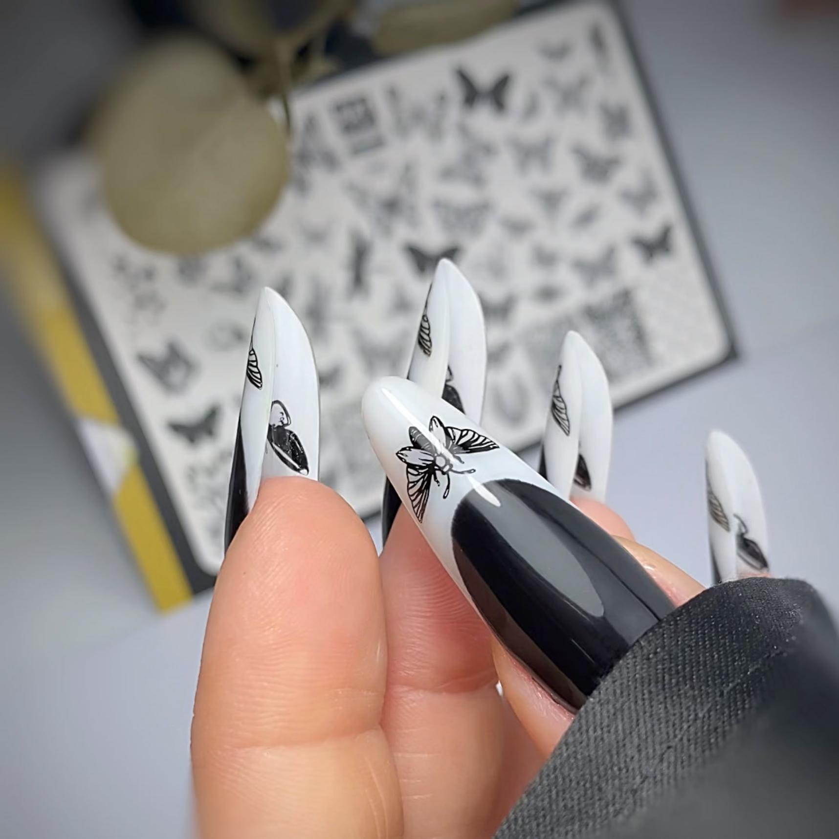 Black and white nail art designs held by a hand with a blurred background of butterfly stamping plates.