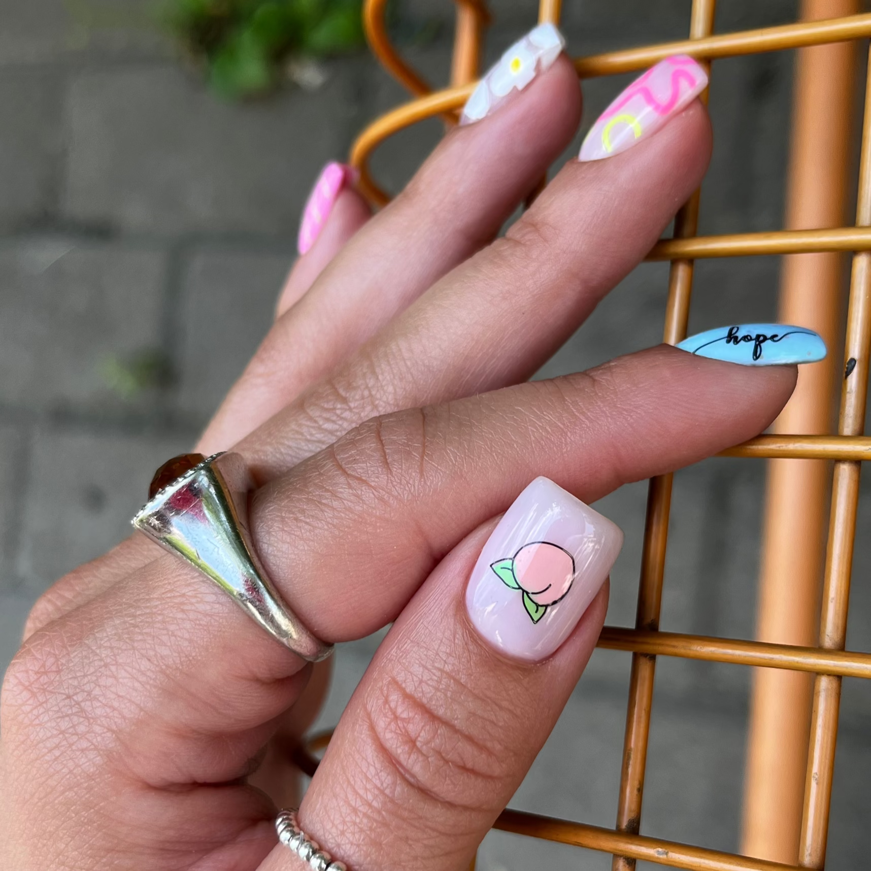 Hand with decorated nails featuring floral and fruit designs, wearing a silver ring, against a blurred background.