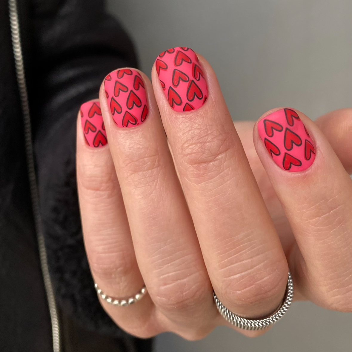 Close-up of a hand with pink nails featuring red heart patterns, wearing silver rings.