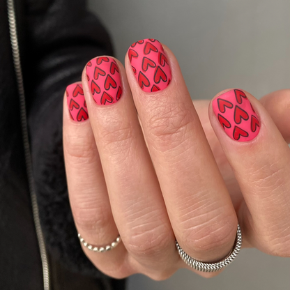 Close-up of a hand with pink nails featuring red heart patterns, wearing silver rings.