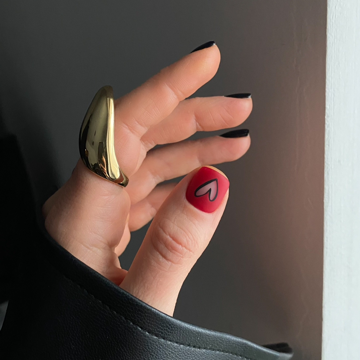 Hand with red nail polish and black shape of hart on that nail and golden ring against a neutral background
