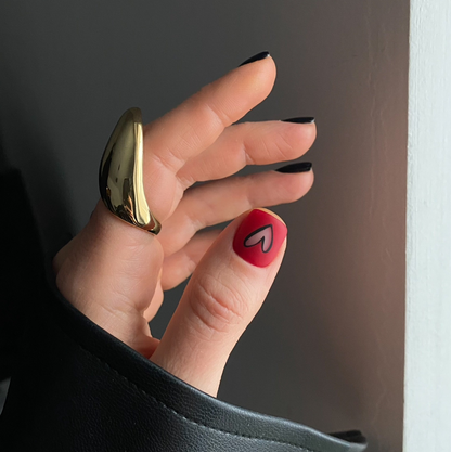 Hand with red nail polish and black shape of hart on that nail and golden ring against a neutral background