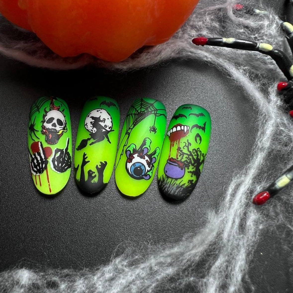 Halloween-themed green nail art with skull, ghost, and pumpkin designs on a dark surface with spider webs.
