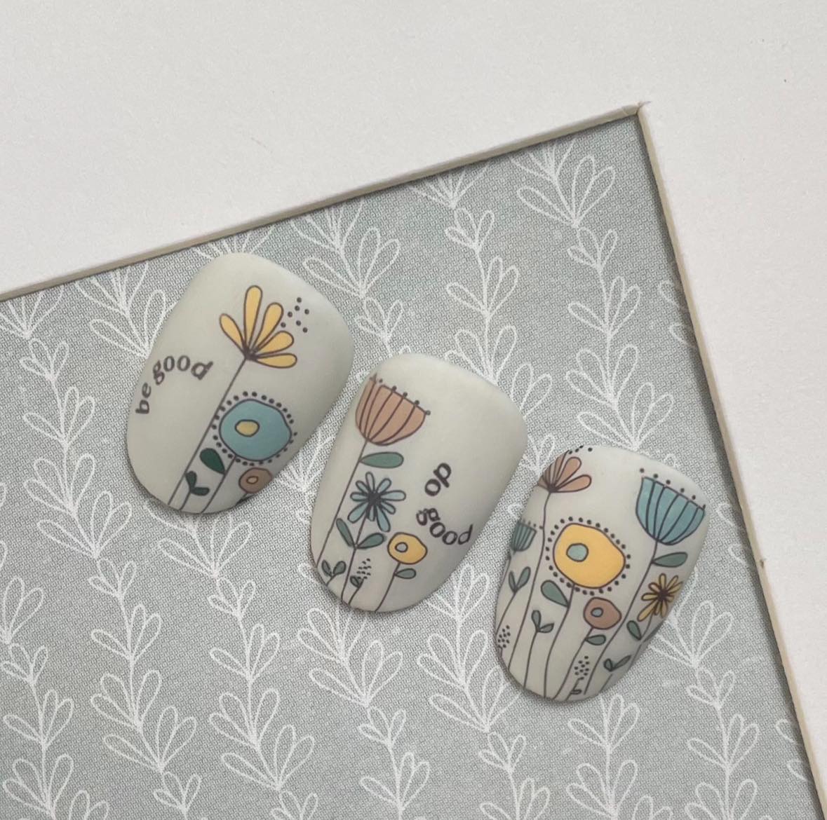 Nails with floral designs and 'be good' text on a gray background with white patterns
