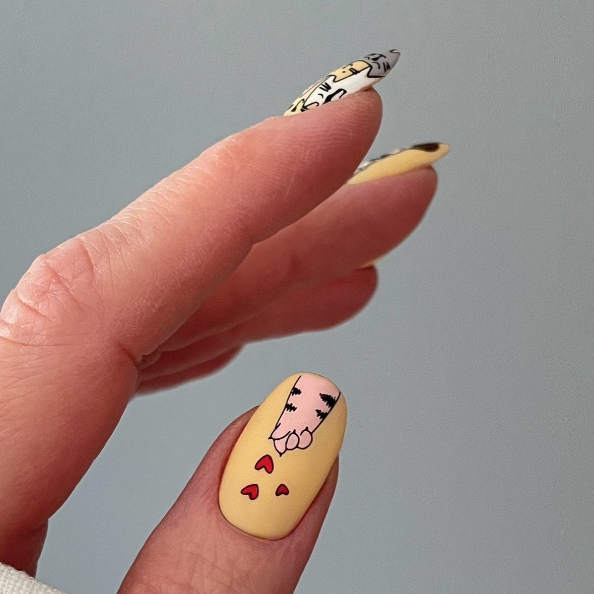 Nail art design on a yellow nail with a face and hearts, held by a hand against a yellow background.
